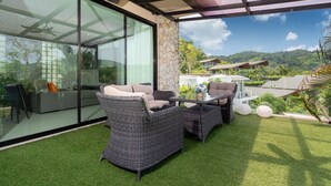 A modern outdoor patio with wicker furniture, a glass wall, and a view of a lush landscape. - Designer Sea View Villa Natural Touch A1 (Kamala)