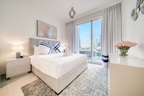 A modern hotel room with a large bed, a nightstand, a bedside lamp, a view of the city through large windows, and a decorative vase with flowers.