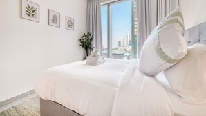 A neatly made bed with white linens, a view of a cityscape through a large window, and three framed botanical prints on the wall.