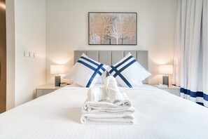 A neatly made bed with white linens and blue pillows, a bedside lamp, and a framed artwork on the wall.