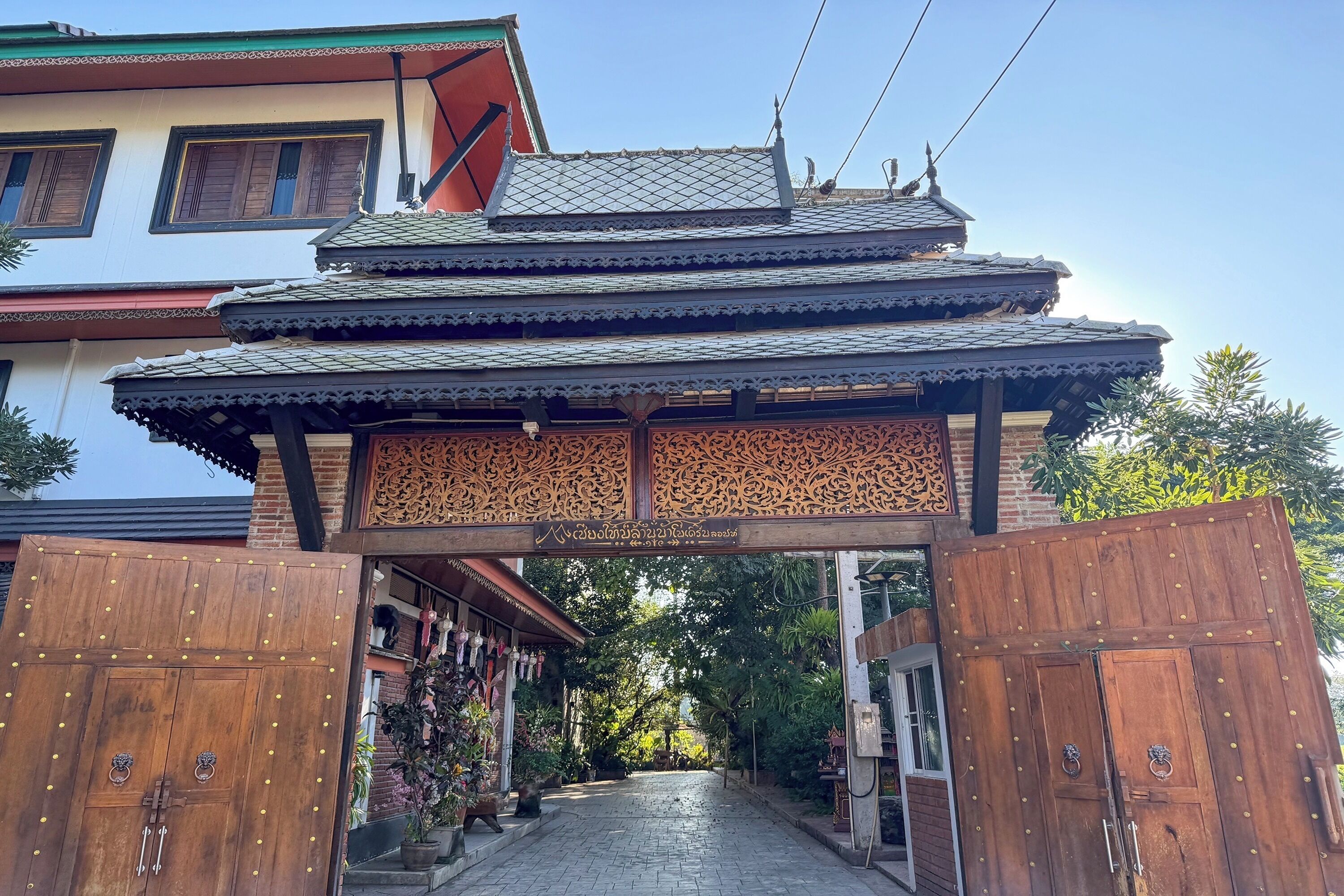 A traditional multi-tiered gate with intricate carvings, leading to a pathway flanked by buildings with balconies and potted plants.