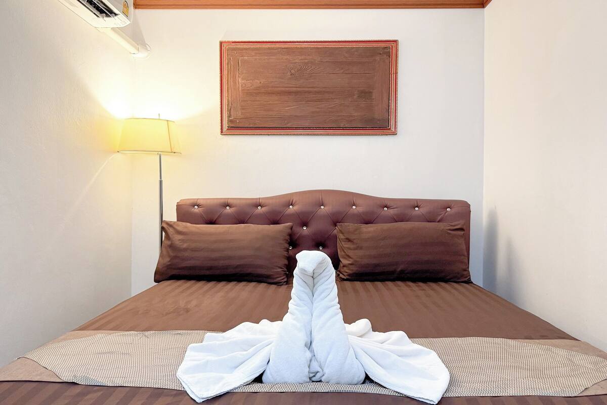 Ein ordentlich bezogenes Bett mit brauner Kopfteil, einer Nachttischlampe und einem gerahmten Bild an der Wand.