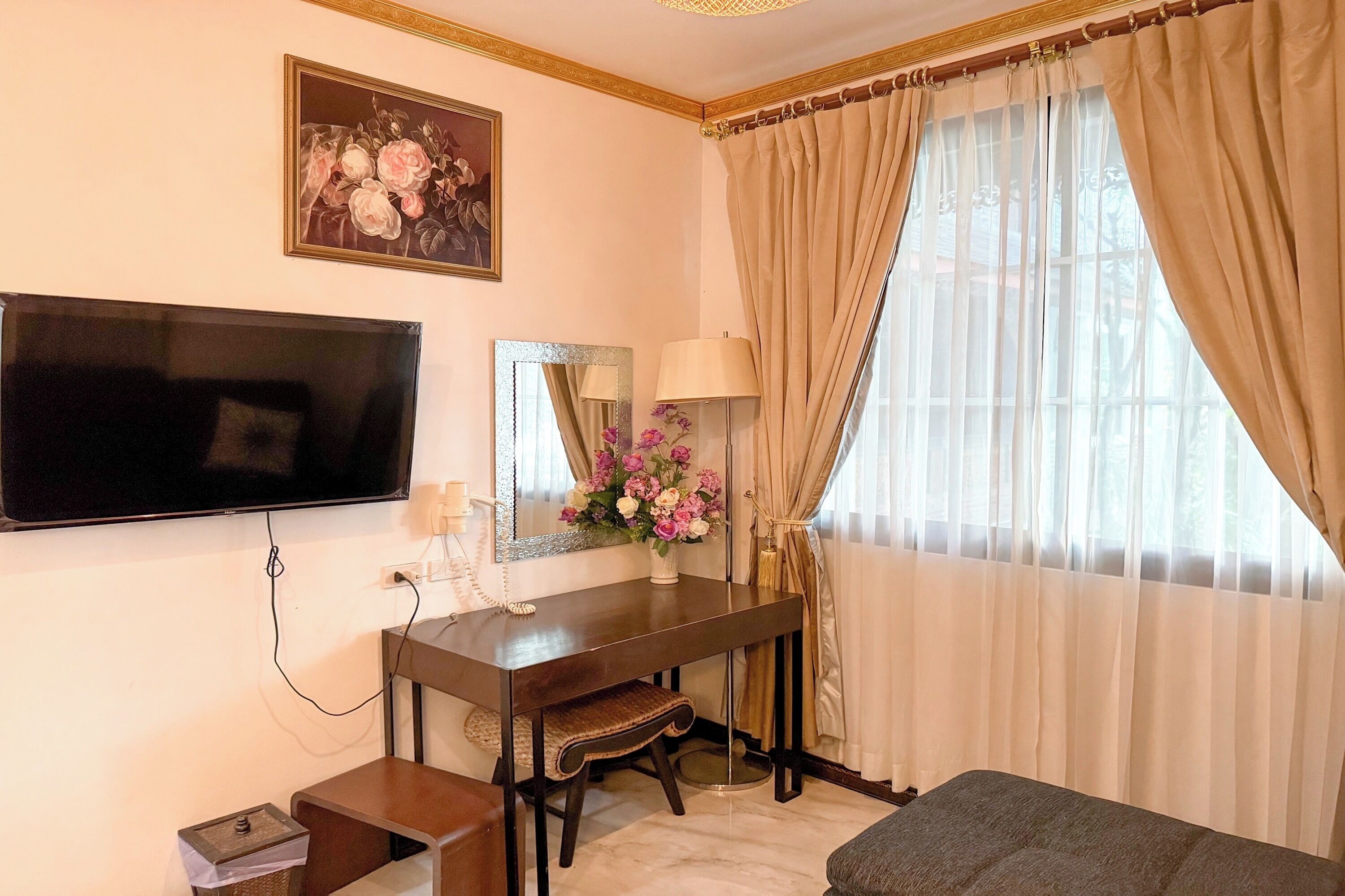 Habitación de hotel con televisor de pantalla plana, un escritorio de madera con lámpara, un espejo y un jarrón de flores.