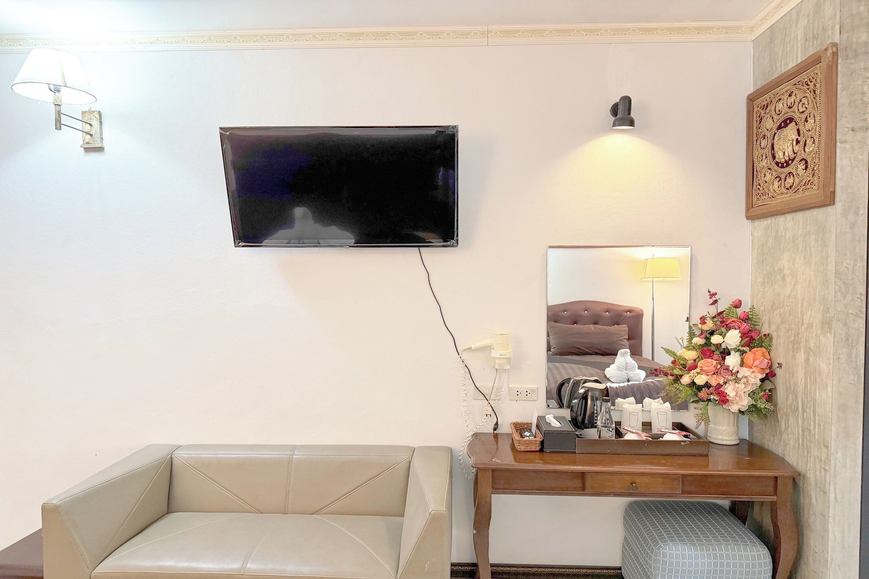 Habitación de hotel con un sofá color crema, un escritorio de madera con un teléfono, un jarrón de flores y un televisor de pantalla plana montado en la pared.