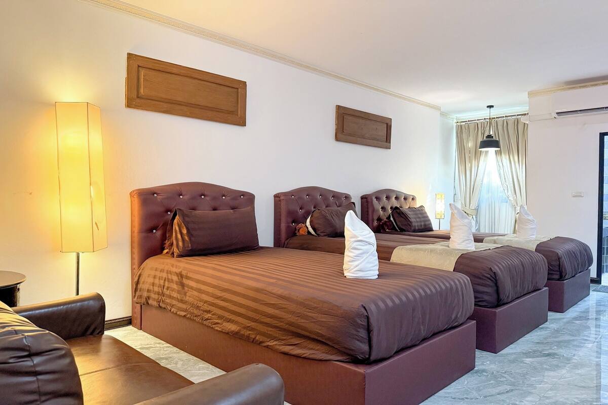 Ein Hotelzimmer mit einem großen Bett, einem kleineren Bett und einem Sofa. Eine Lampe, ein Fenster mit Vorhängen und eine Wandklimaanlage sind vorhanden.