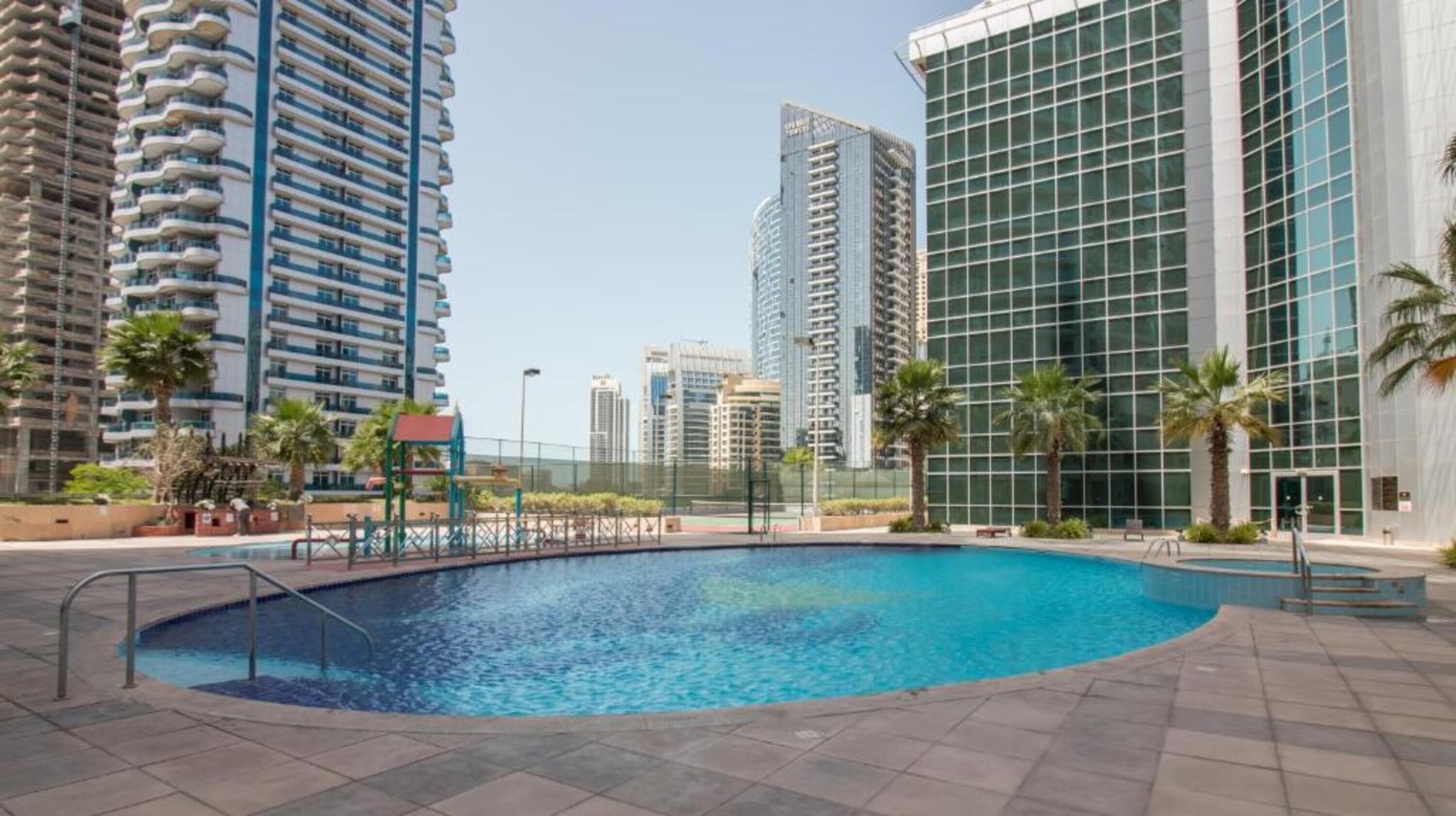A swimming pool surrounded by high-rise buildings, with palm trees and a fenced area.