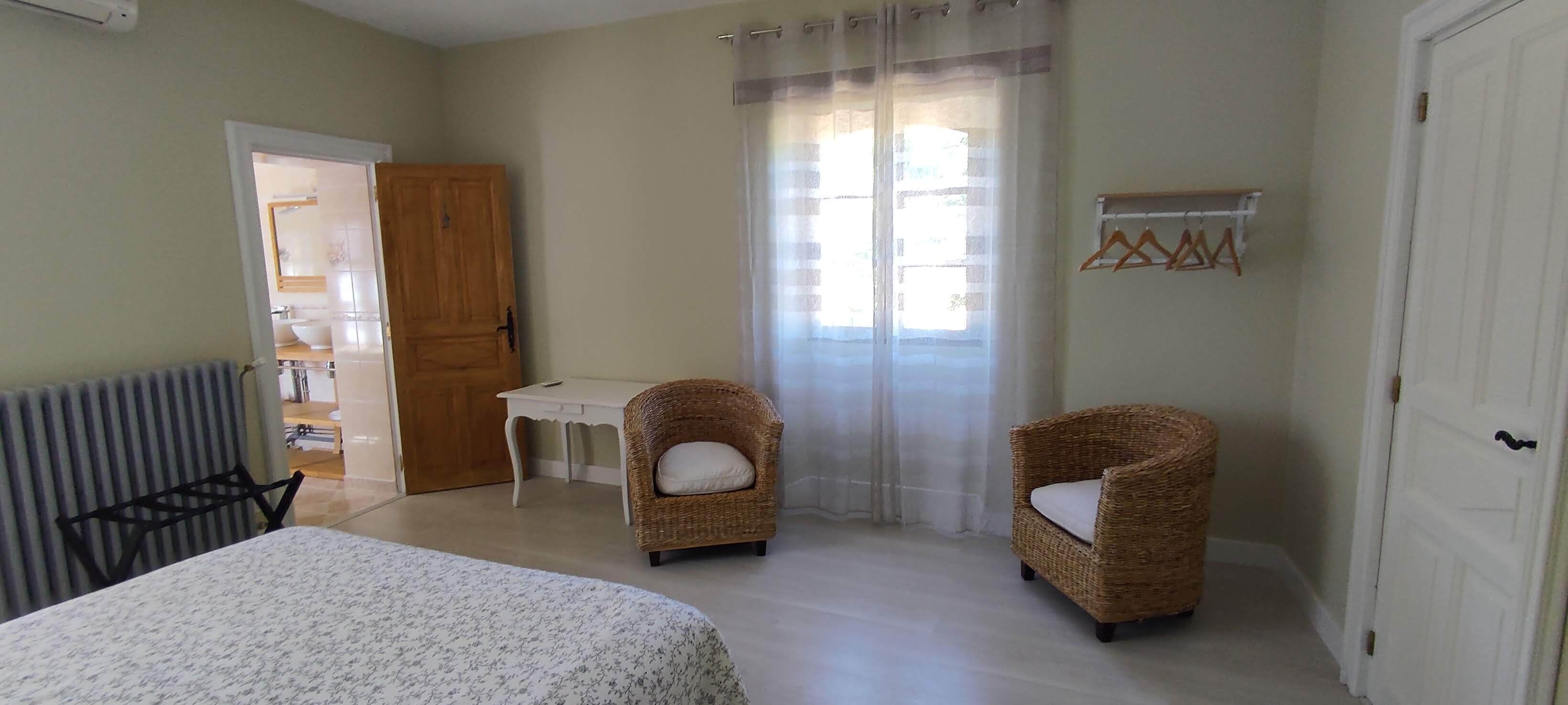 A bedroom with a bed, two wicker chairs, a small table, a radiator, a wooden door, and a window with curtains.