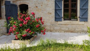 A stone building with a window and shutters, a flower bed with pink flowers, and a stone pathway.