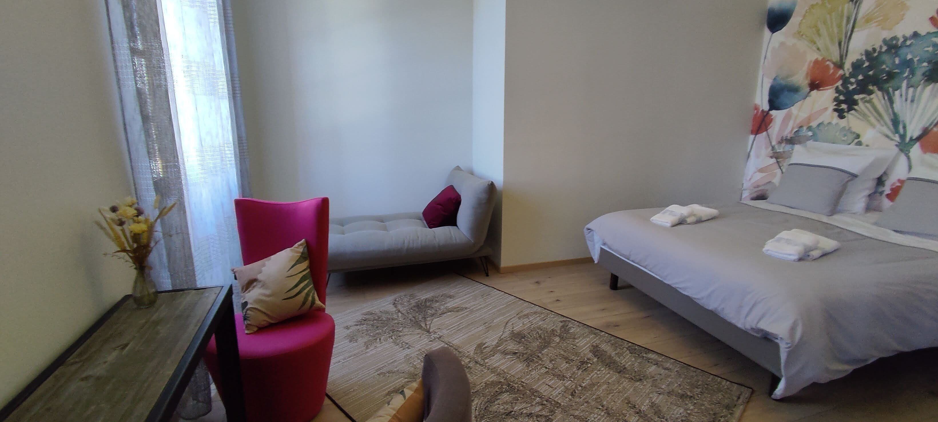 A bedroom with a bed, a red chair, a grey sofa, and a wooden table.