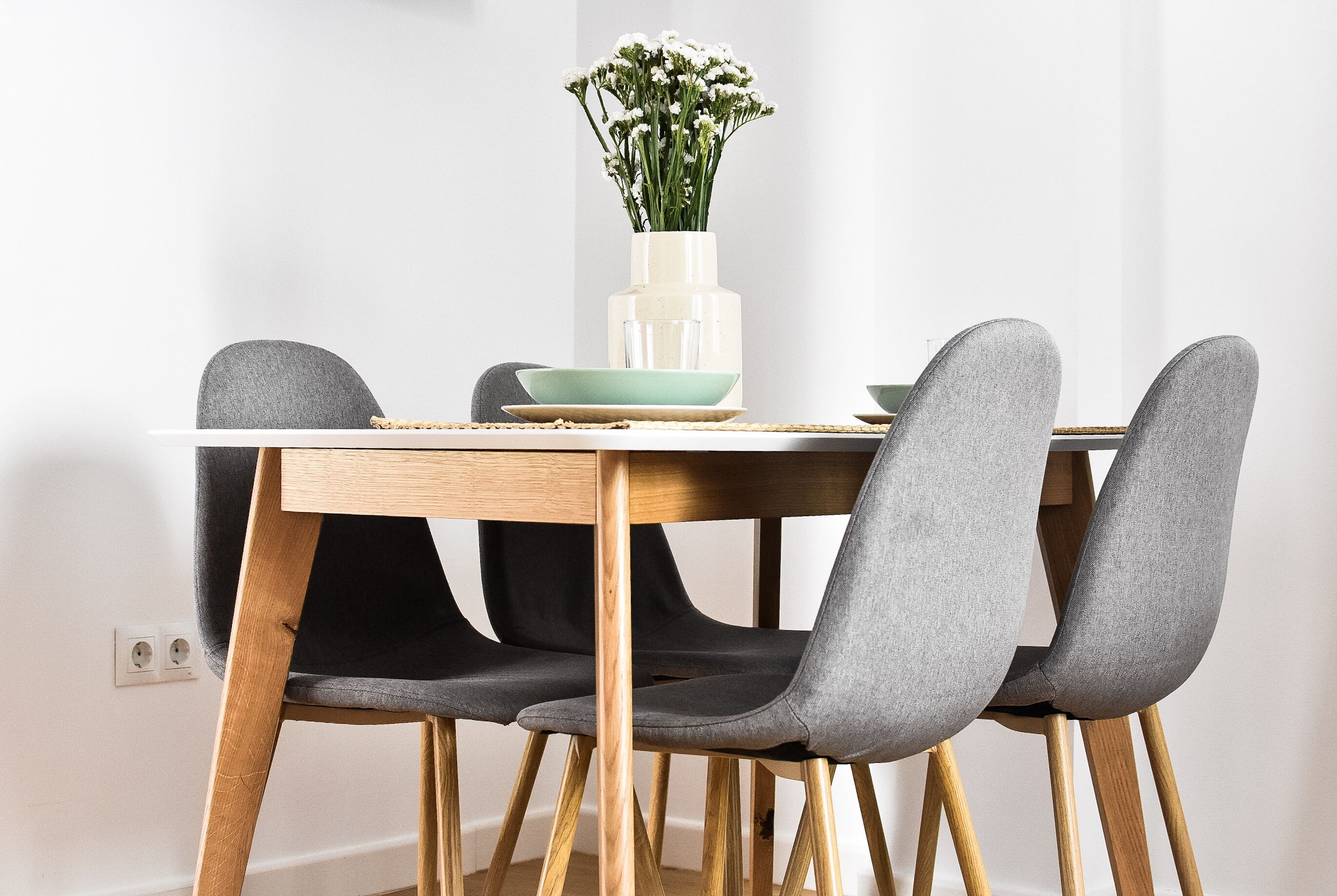 A dining table with four grey upholstered chairs, a white vase with flowers, and a green bowl.