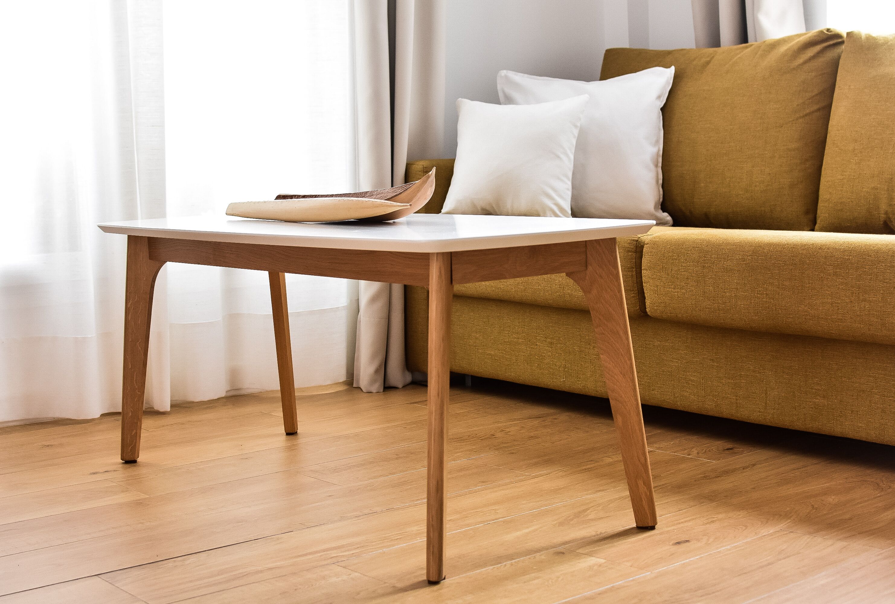 Ein Holztisch mit weißer Oberfläche und einem braunen Gegenstand darauf, vor einer senffarbenen Couch mit weißen Kissen platziert.