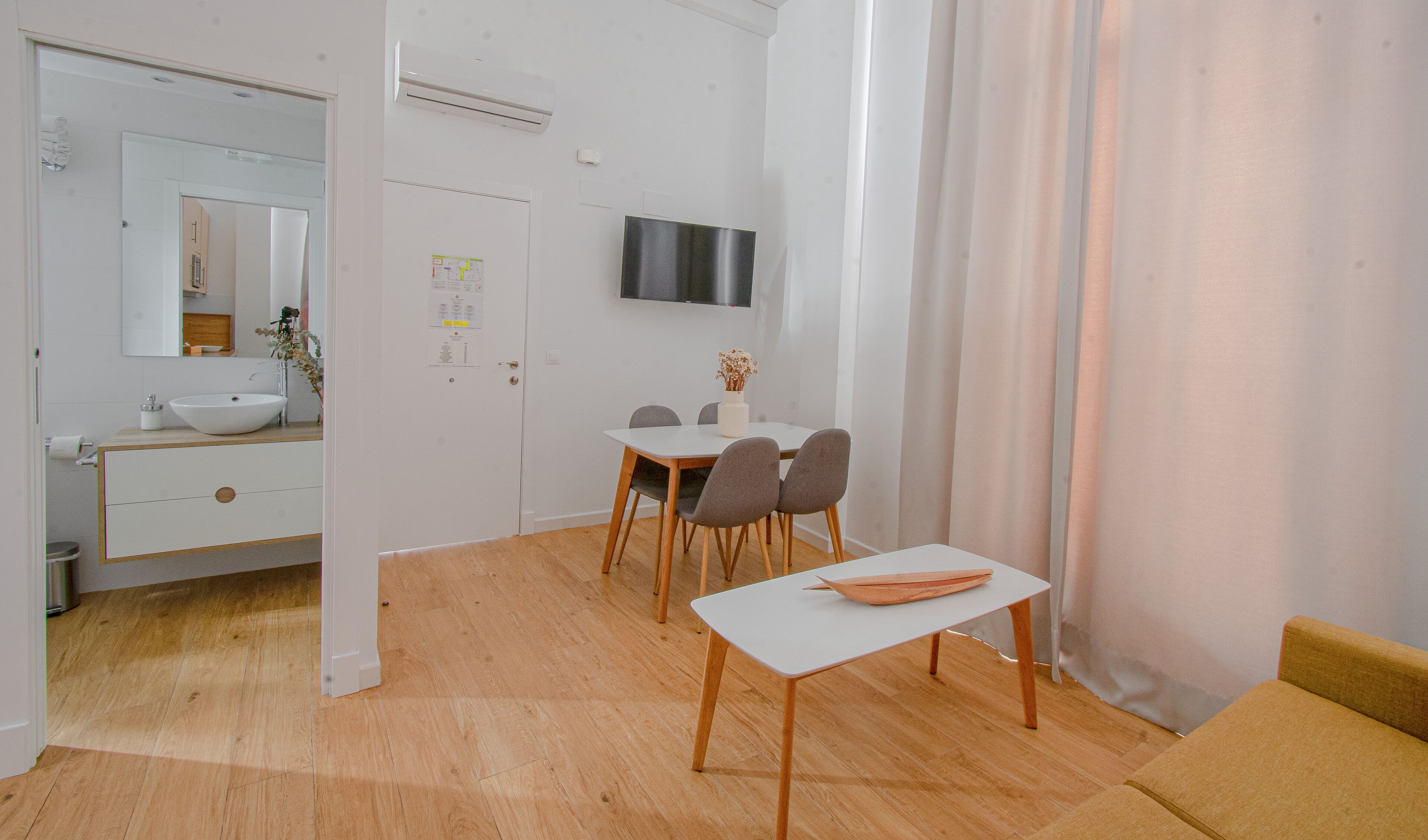 Une salle de bain moderne avec un lavabo blanc, un miroir et des toilettes. On y trouve également un coin repas avec une table et des chaises, ainsi qu’une petite table sur laquelle est posé un vase.