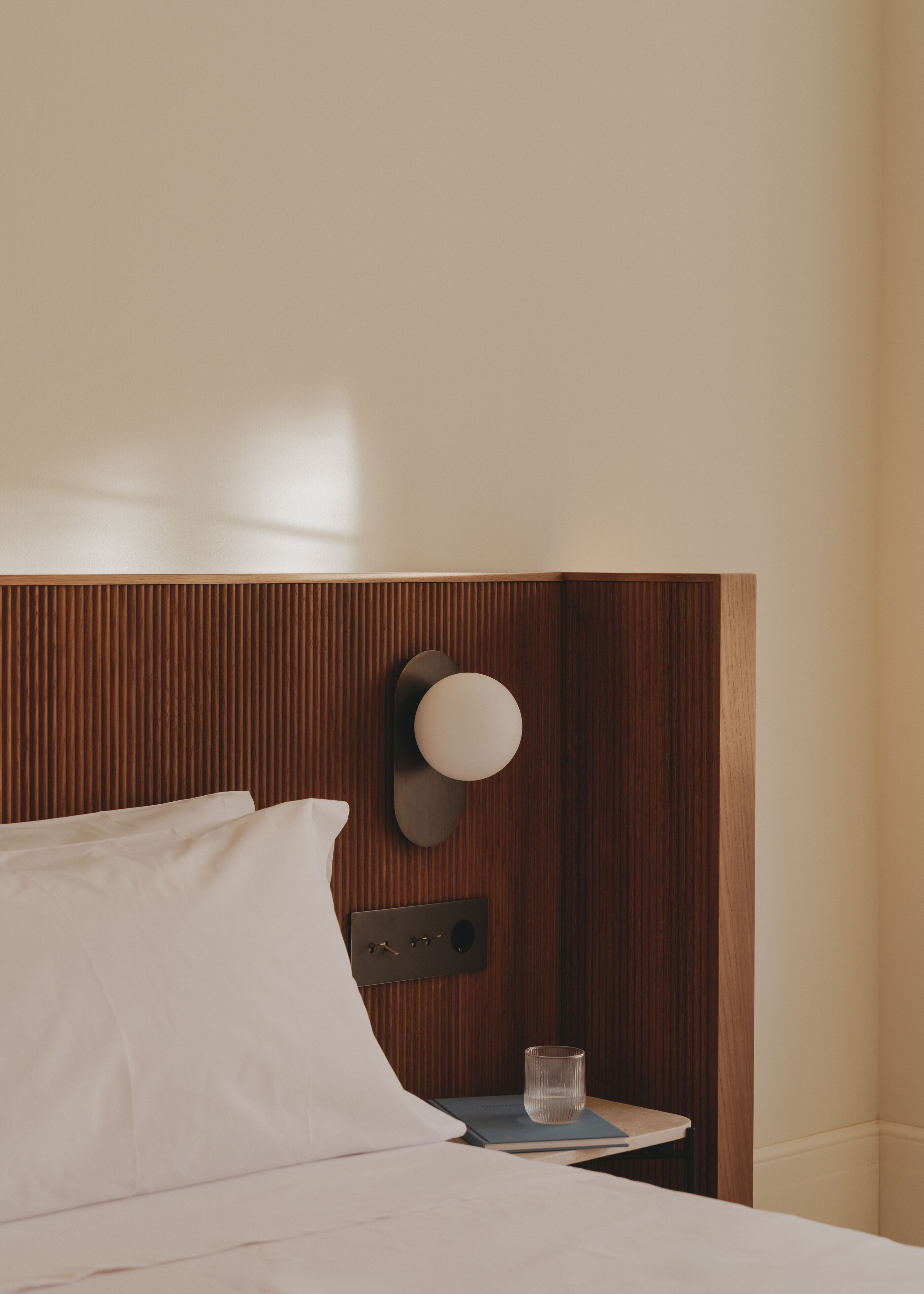 A neatly made bed with white linens, a bedside table with a glass of water, and a wall-mounted light fixture.