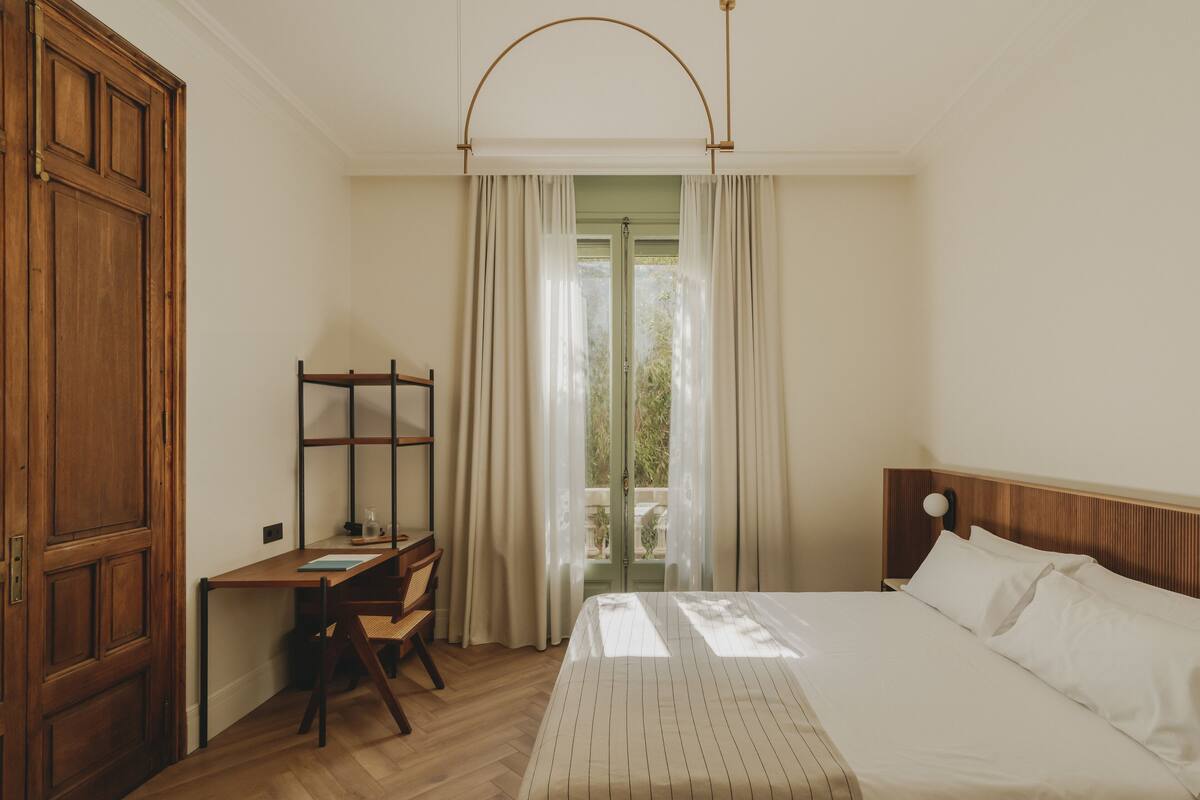 A bedroom with a wooden desk, a bed with white linens, and a view of greenery through a window with curtains.