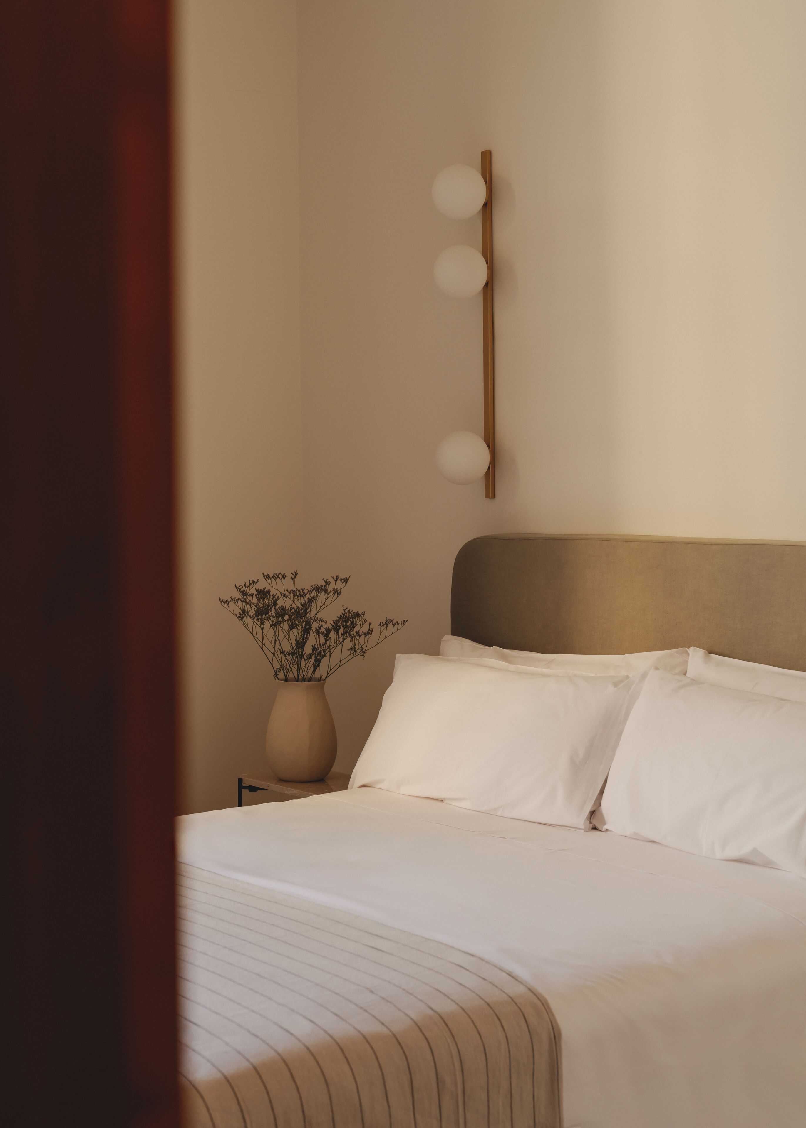 Cama bem arrumada com lençóis brancos e cabeceira, mesa de cabeceira com um vaso de flores, e luminária fixada na parede.