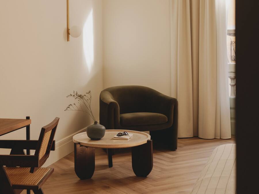 A minimalist living room with a wooden floor, a dark brown armchair, a round coffee table, and a dining area with a wooden table and chairs.