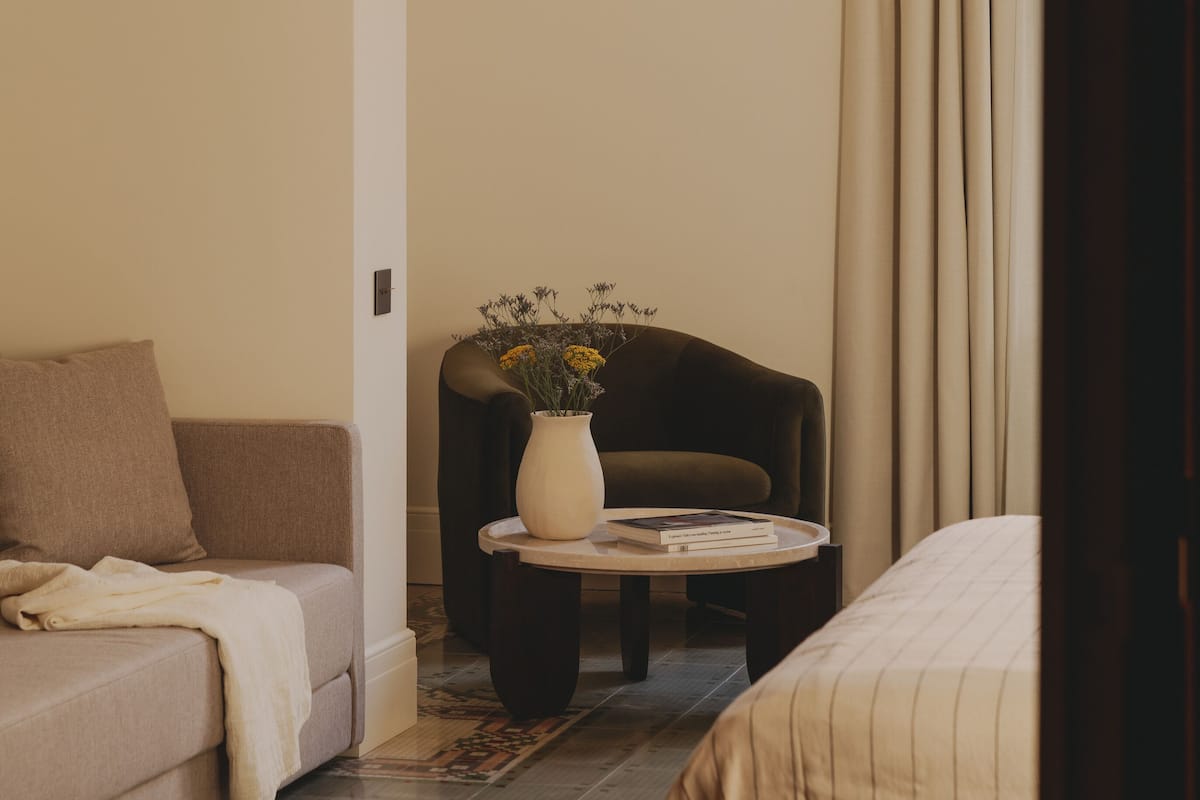 A hotel room with a sofa, a chair, a small round table with a vase of flowers, and a bed.
