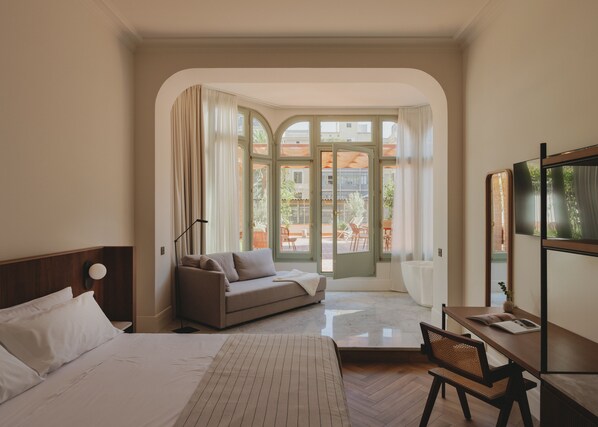 Una habitación de hotel con cama, sofá, escritorio y un amplio ventanal con cortinas.