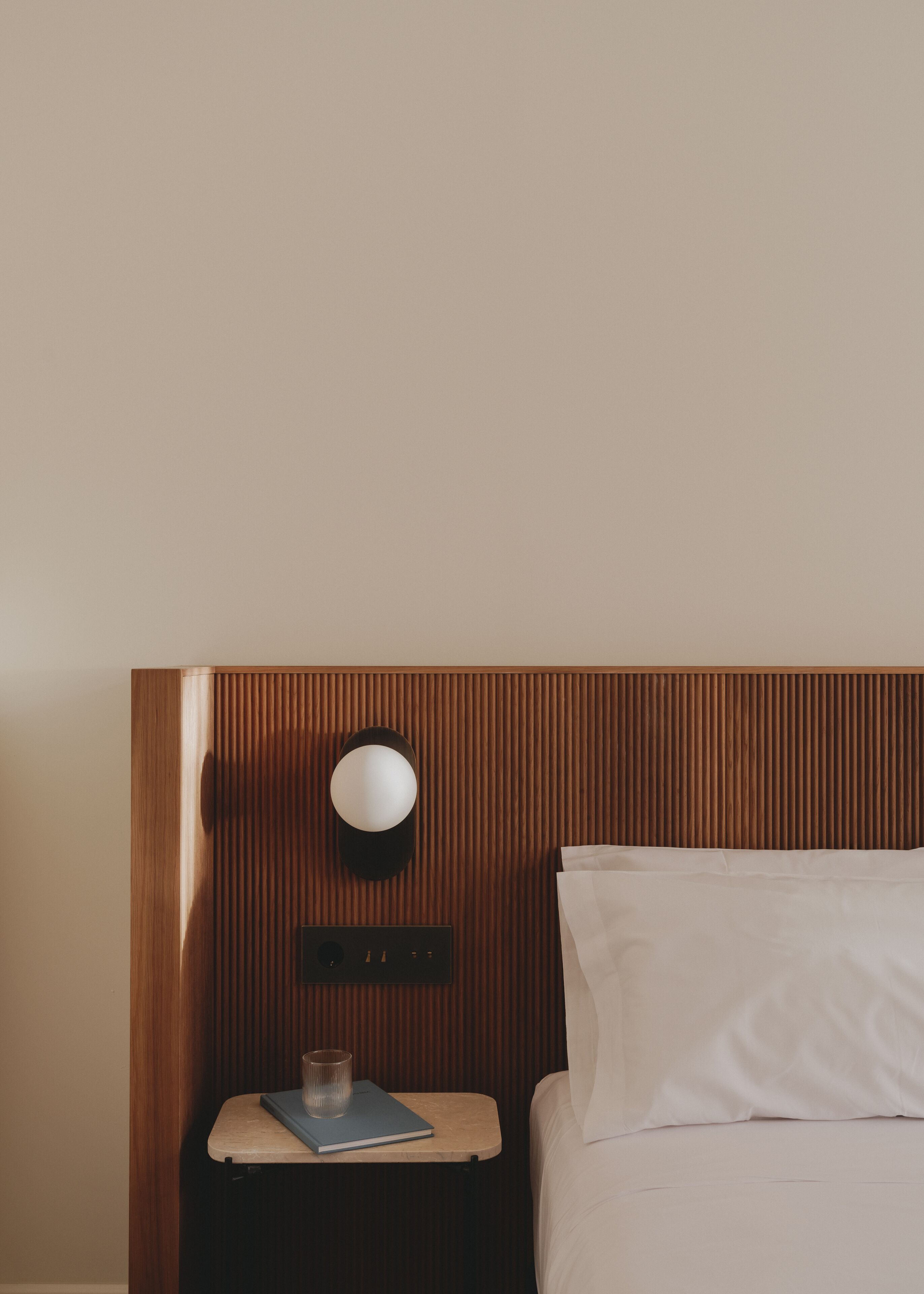 Habitación de hotel con cabecera de madera, mesita de noche con lámpara, un libro y un vaso.