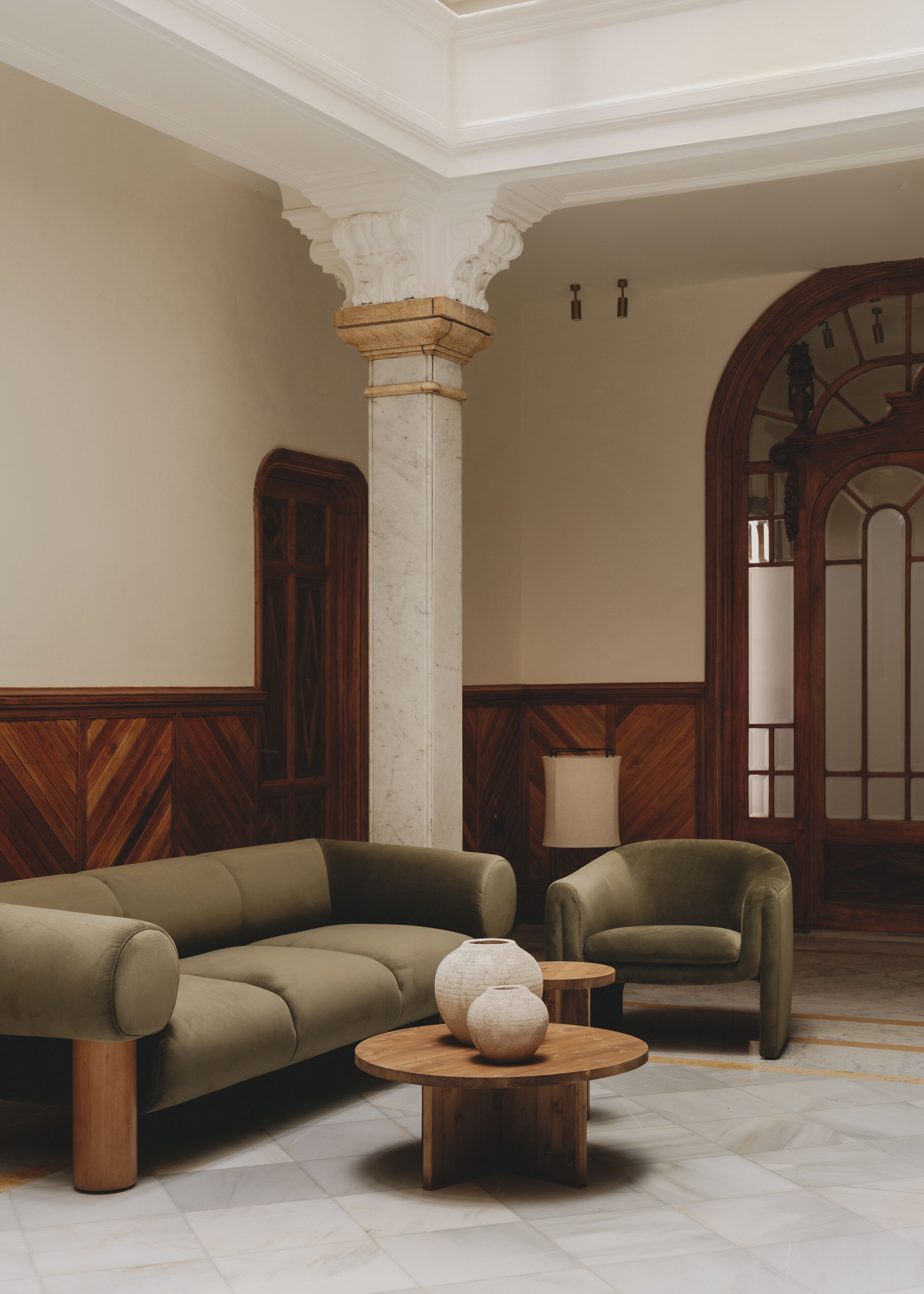 A room with a large wooden cabinet, a beige sofa, a smaller armchair, and a round wooden coffee table with two white vases.