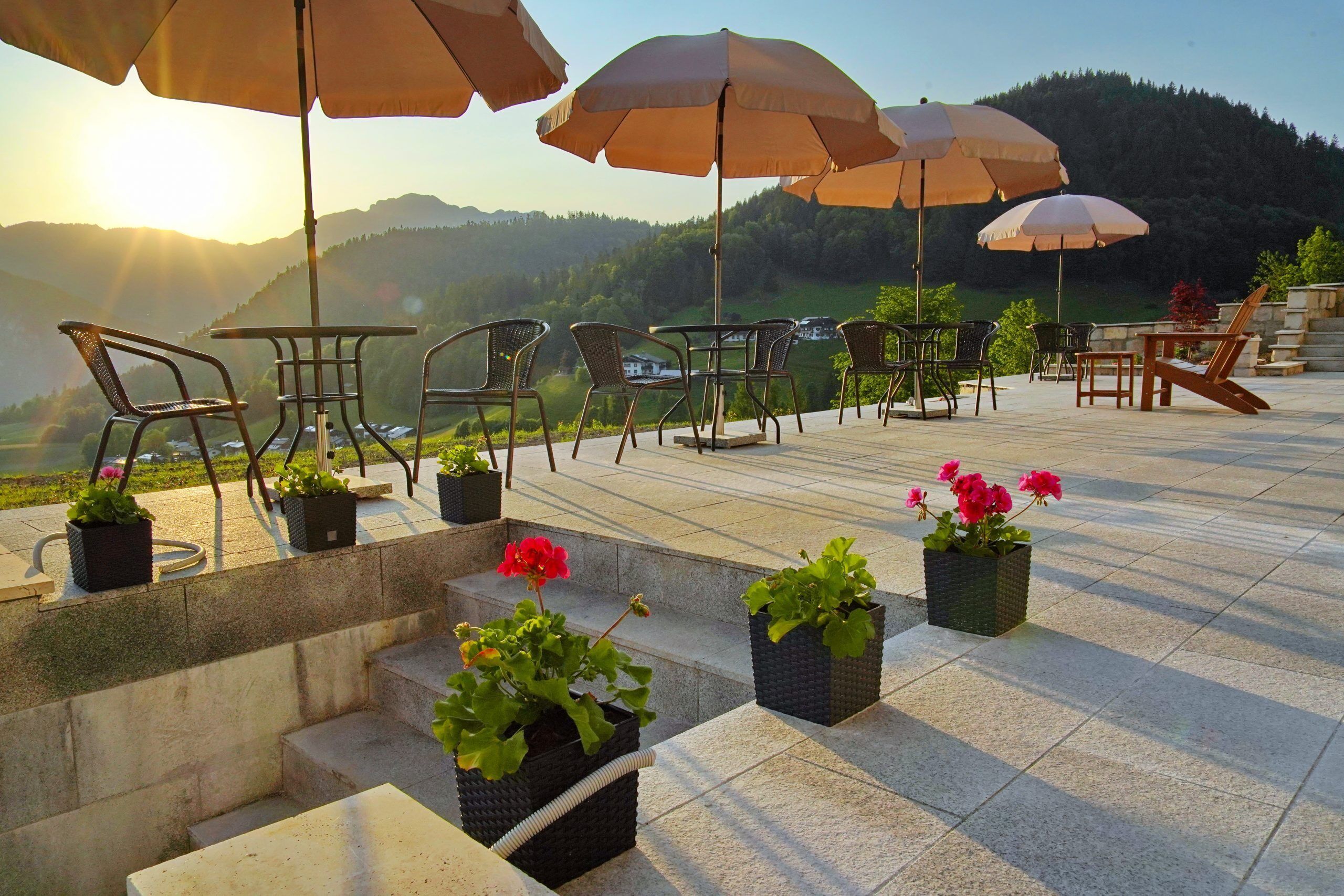Terraza al aire libre con mesas y sillas, rodeada de plantas en macetas y sombrillas, con vistas a un paisaje pintoresco.