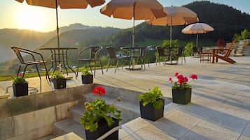 Terraza al aire libre con mesas y sillas, rodeada de plantas en macetas y sombrillas, con vistas a un paisaje pintoresco.