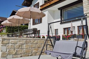 A patio area with outdoor seating, including a swing chair and tables, under large umbrellas, with a stone wall and flower boxes in front of a building. - Villa Bello (Berchtesgaden)
