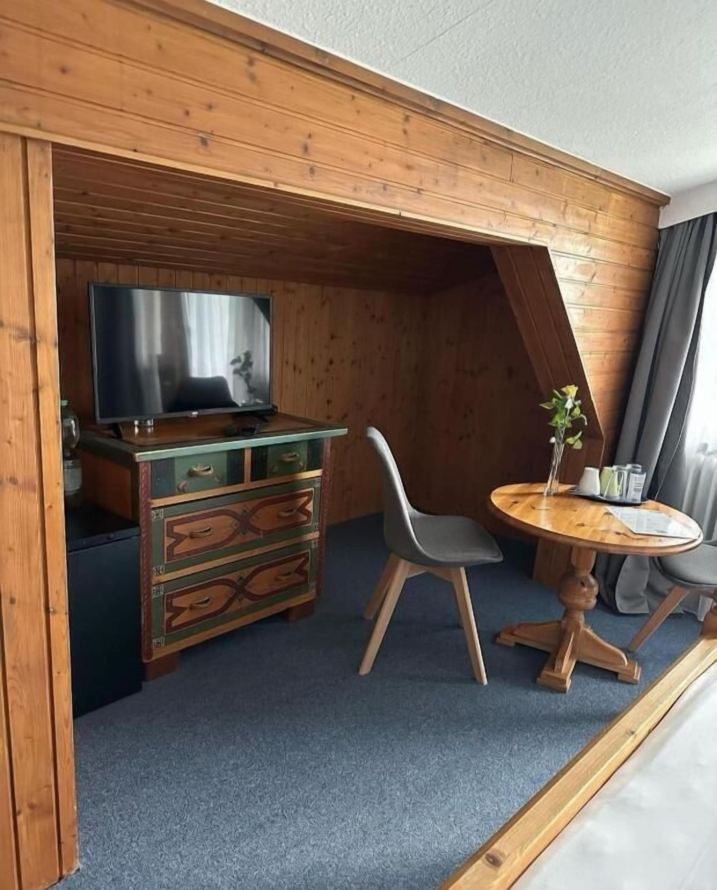A cozy room with a wooden ceiling, a TV on a wooden stand, a small wooden dresser, two grey chairs, and a round wooden table.