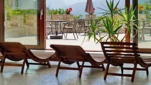 An indoor pool area with lounge chairs and a table, large windows with a mountain view, and a ceiling with wooden slats. - Villa Bello (Berchtesgaden)