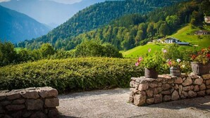 A stone wall with potted plants and a view of a house on a hill with lush greenery and mountains in the background. - Villa Bello (Berchtesgaden)