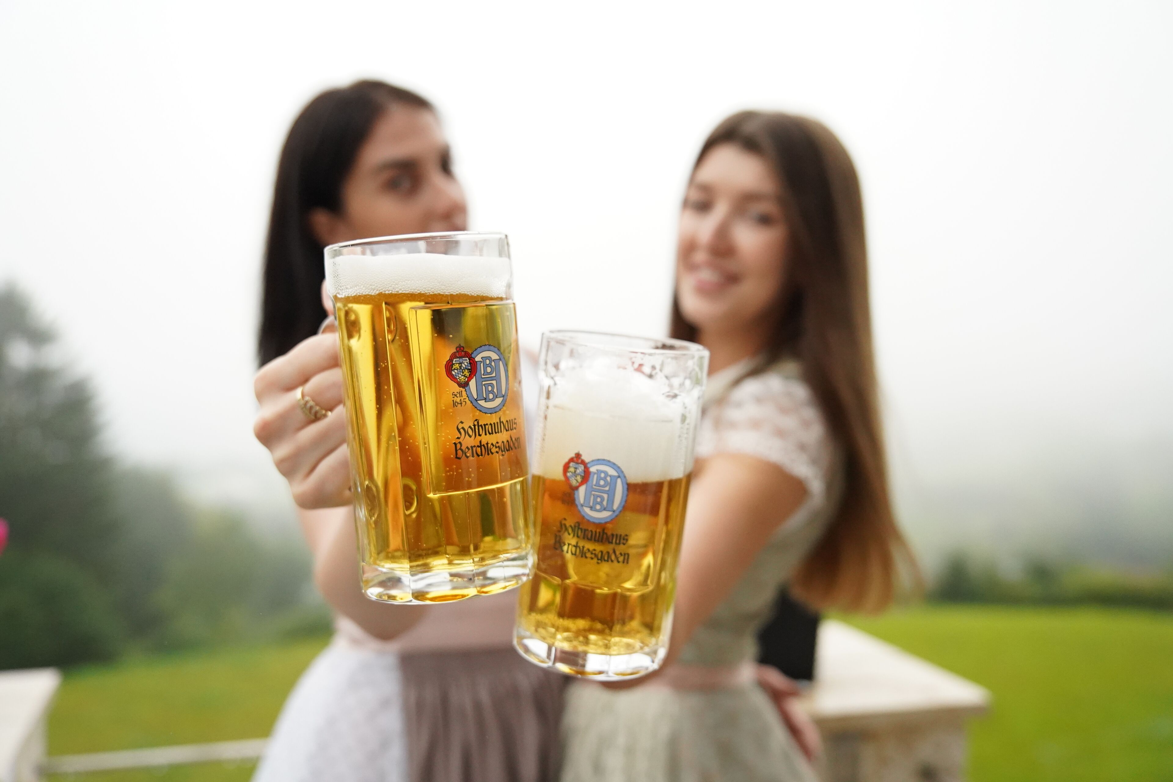 Twee personen met bierglazen met een logo, buiten met een grasveld op de achtergrond.