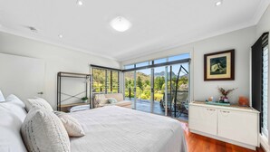 A bedroom with a large bed, a view of the outdoors, a dining area, and a white cabinet with decorative items.