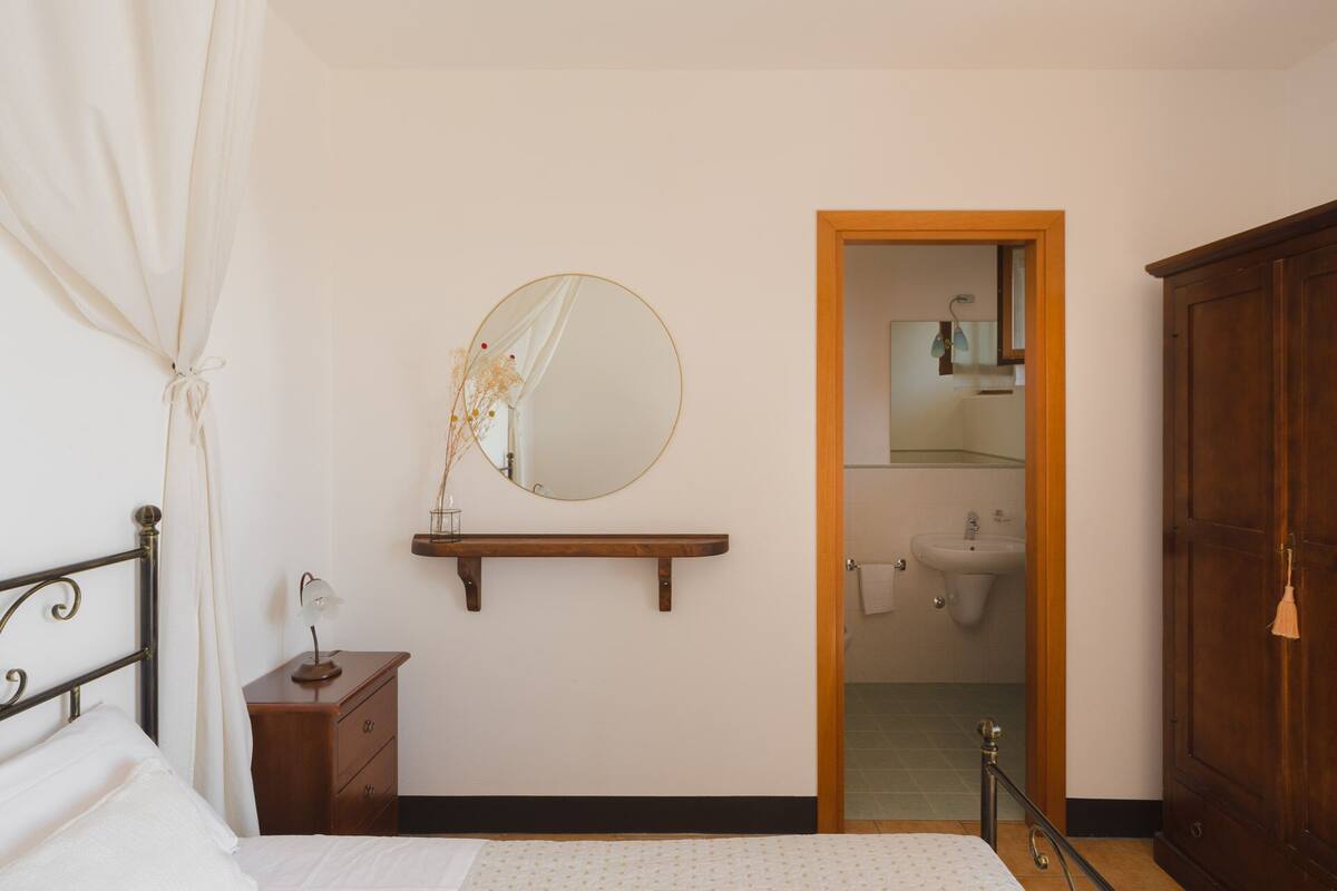 Un dormitorio con cama, una mesita de noche con lámpara, un banco de madera, un espejo y un baño visible a través de una puerta abierta.