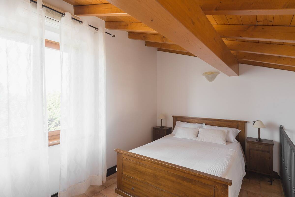 Un dormitorio con una cama de madera, dos mesitas de noche con lámparas, una ventana con cortinas traslúcidas y un techo inclinado.