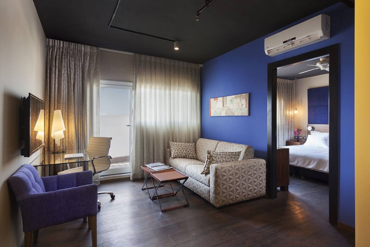 Una camera d'albergo moderna con divano, poltrona e un tavolino. Si nota un letto con testiera e un comodino con una lampada. La stanza dispone di un'ampia finestra con tende, un condizionatore d'aria e un quadro appeso al muro.