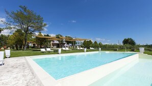 Una piscina rettangolare con poltroncine bianche e un'area verdeggiante con alberi e un edificio sullo sfondo.