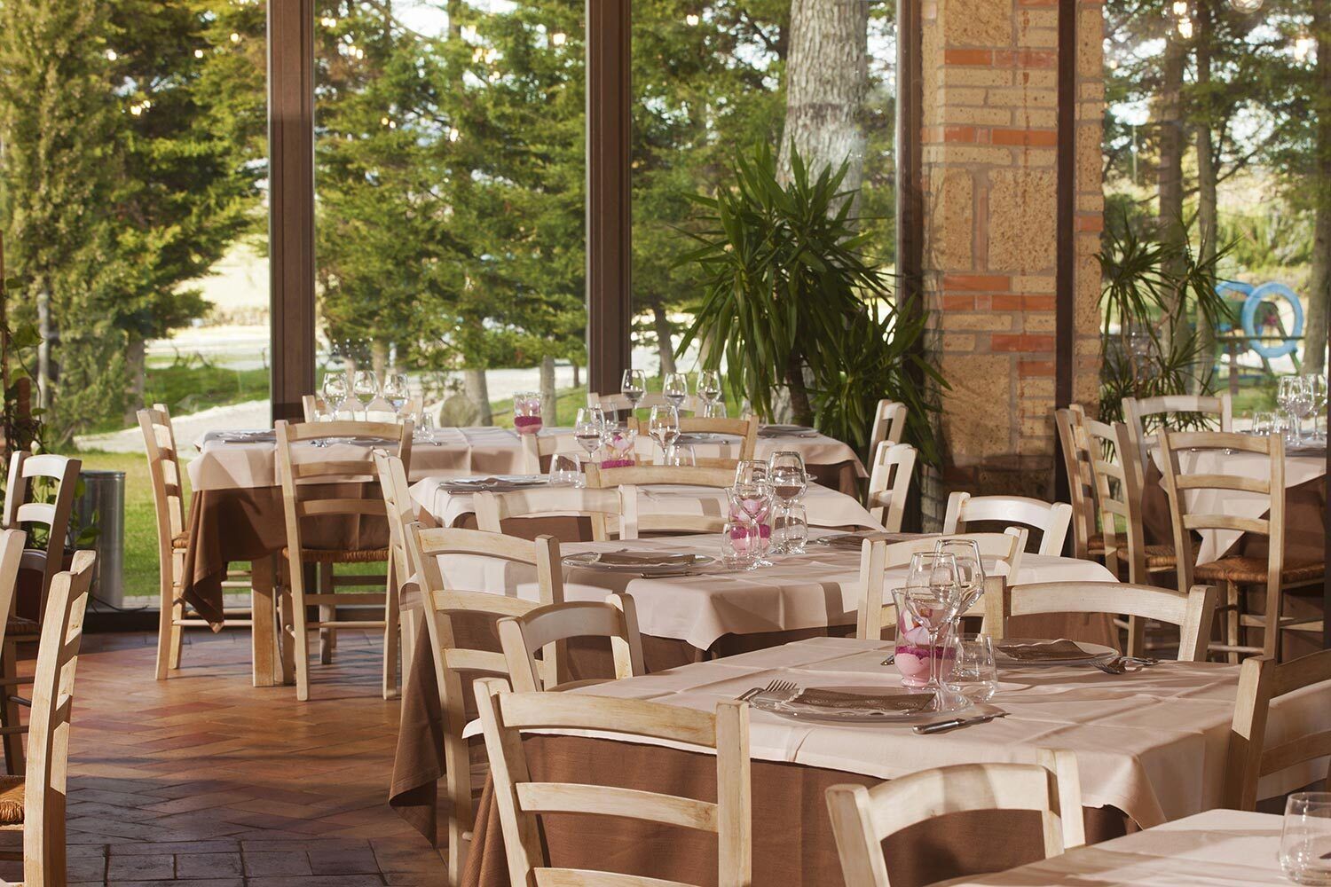 An outdoor dining area with multiple tables and chairs, set against a backdrop of trees and a brick wall.