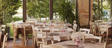 An outdoor dining area with multiple tables and chairs, set against a backdrop of trees and a brick wall.