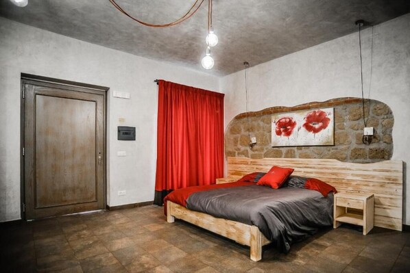 A bedroom with a wooden bed, red curtains, a stone wall, and a red flower painting. - Agriturismo La Collina (Pitigliano)