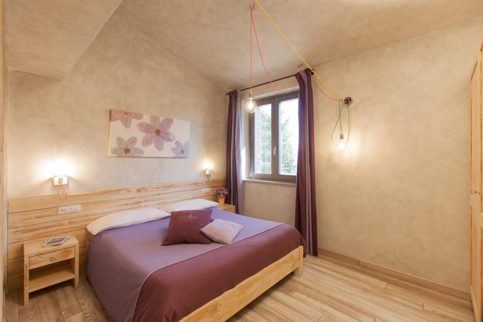 A bedroom with a wooden headboard, a bed with a purple bedspread, a nightstand with a lamp, a window with curtains, and a wall-mounted light fixture.