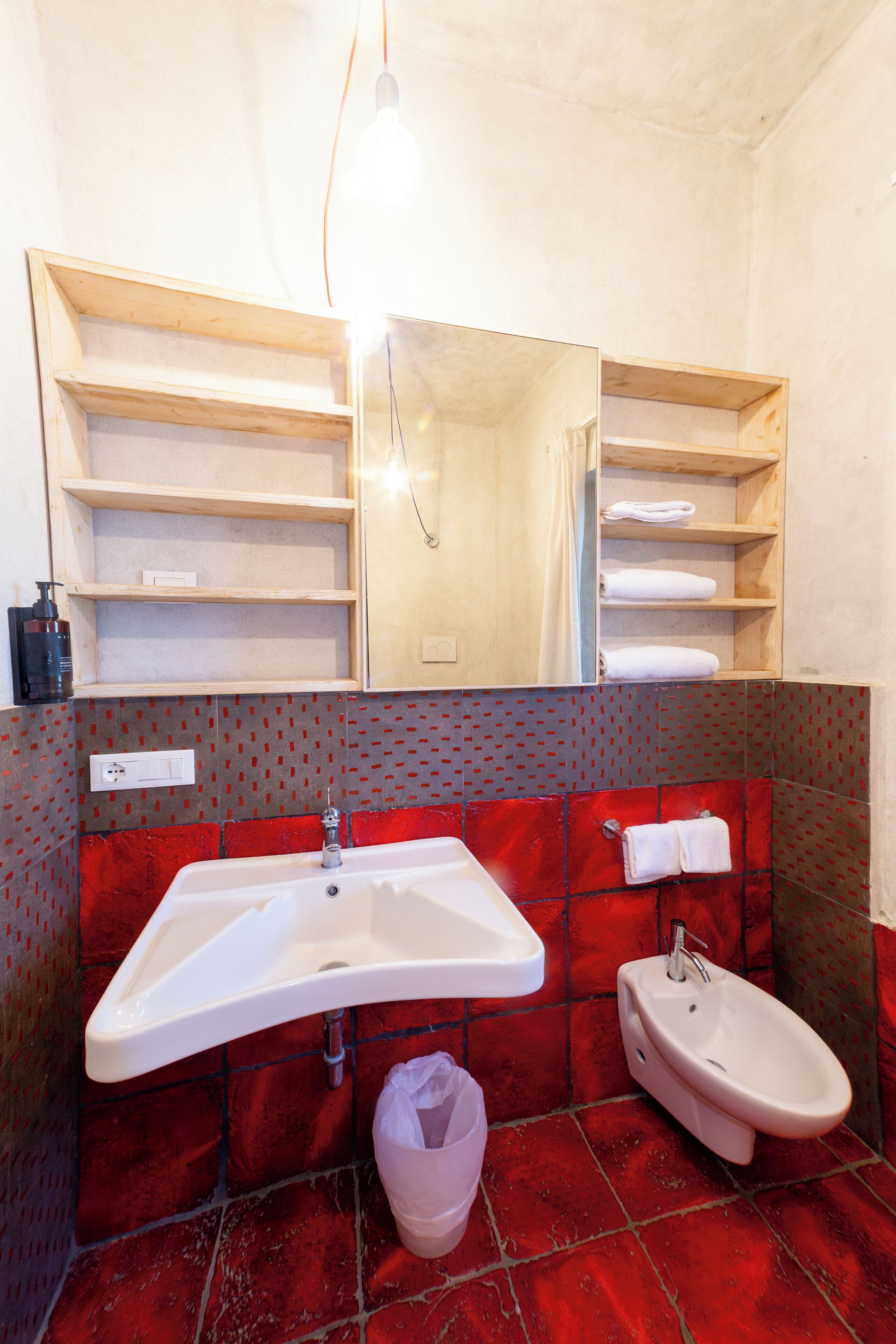 Ein Badezimmer mit einem weißen Waschbecken, einem Bidet und einem Spiegel darüber. Über dem Waschbecken befinden sich Holzregale und darunter ein Mülleimer.