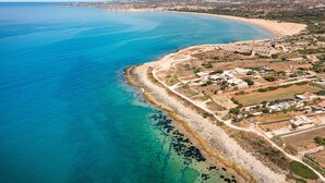 An aerial view of a coastal area with a beach, clear blue waters, and a mix of residential and agricultural land. - Stunning beachfront villa with pool and direct access to the sea in Sampieri - DiCasaInSicilia (Sampieri)