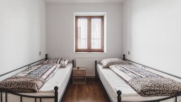 A dormitory room with two single beds, a small wooden nightstand, and a window with blinds.