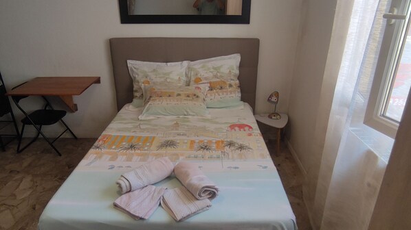 A bed with a patterned bedspread, pillows, and rolled towels. There is a small table and a chair to the left of the bed.