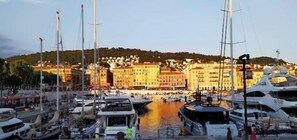 A marina with multiple yachts docked, a waterfront building complex in the background, and a hilly landscape.