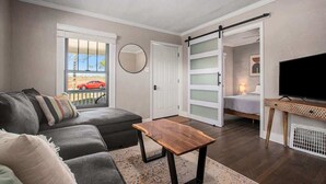 Ein Wohnzimmer mit einem grauen Sofa, einem Holztisch, einem Fernseher auf einem Schrank und einer Schiebetür, die in ein Schlafzimmer führt.