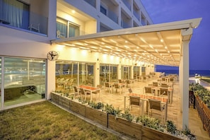 A covered outdoor dining area with tables and chairs, a view of the sea, and a building with large windows. - GADILEH RESORT HOTEL (Tadjoura)