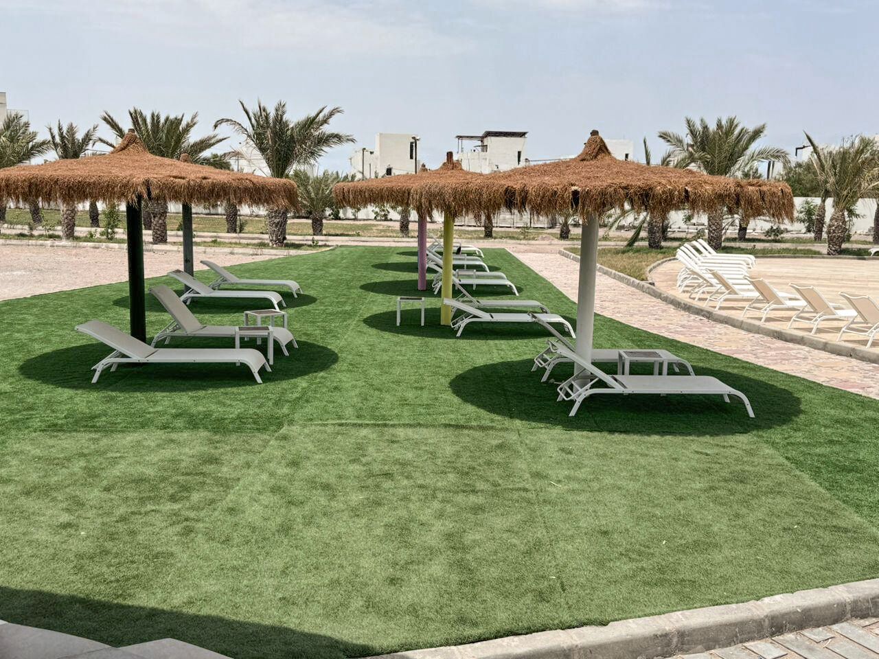 A pool area with sun loungers and thatched umbrellas, surrounded by palm trees and buildings.