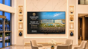 En modern hotellobby med en stor skärm som visar resortens namn och bilder av omgivningarna, bekväma sittplatser och dekorativa vaser.