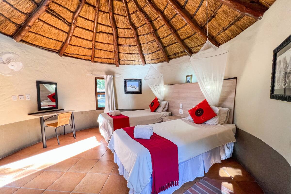 A room with two beds, a desk, a chair, a window, and a ceiling with a thatched roof.